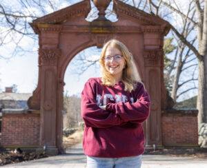 A student wearing a maroon RCNJ sweatshirt is smiling with her arms crossed while standing in front of the Havemeyer Arch. The trees are bare and the sky is blue with streaks of clouds.