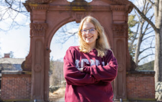 A student wearing a maroon RCNJ sweatshirt is smiling with her arms crossed while standing in front of the Havemeyer Arch. The trees are bare and the sky is blue with streaks of clouds.