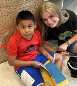 A young woman sits on the floor with a second grade boy. She smiles. He holds a paper.