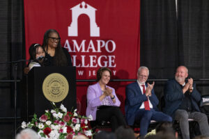 Three people sit in chairs on stage. To the left is a podium, with a young woman behind it. She is being hugged by a woman. They are all in front of a Ramapo College banner.