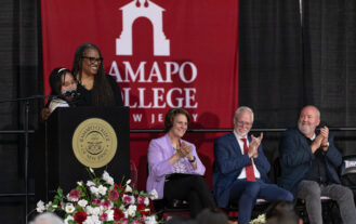 Three people sit in chairs on stage. To the left is a podium, with a young woman behind it. She is being hugged by a woman. They are all in front of a Ramapo College banner.