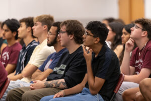 A group of college students sit in chairs and listen. They are in a crowd in a room.