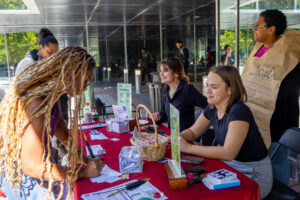 Two people sit behind a table covered in information about voting. A person dressed as the constitution stands behind them. Another person stands up on the other side of the table and fills out a paper.