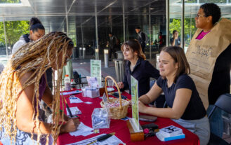 Two people sit behind a table covered in information about voting. A person dressed as the constitution stands behind them. Another person stands up on the other side of the table and fills out a paper.