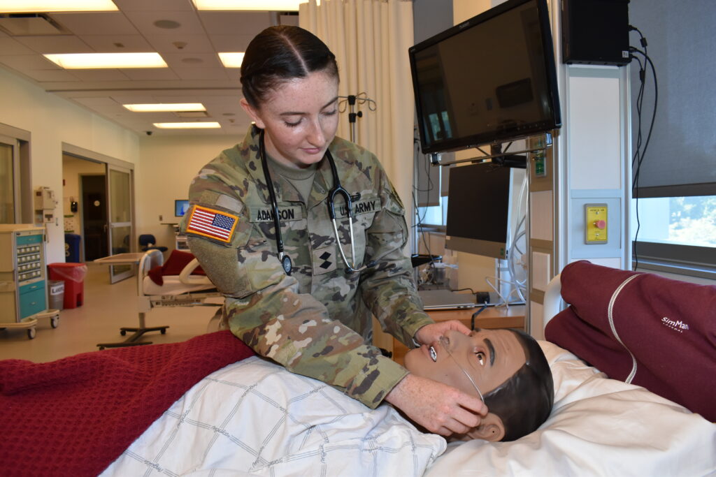 Olivia is dressed in army fatigues and at bedside in the Ramapo College simulation lab checking on a robotic patient