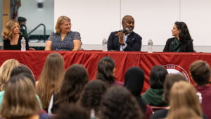 A group of four people sit at a table for a panel discussion. They are in front of a crowd.
