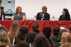 A group of four people sit at a table for a panel discussion. They are in front of a crowd.