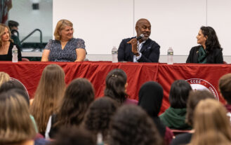 A group of four people sit at a table for a panel discussion. They are in front of a crowd.