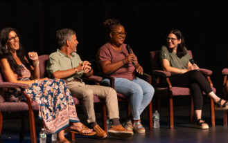 A group of four adults sit on chairs on a stage. One in hold a microphone. They are all smiling and laughing.