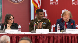 A group of three people sit at a table. The one in the middle has a military uniform on. There is an American flag behind them.