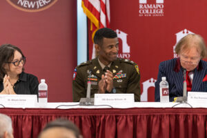 A group of three people sit at a table. The one in the middle has a military uniform on. There is an American flag behind them.
