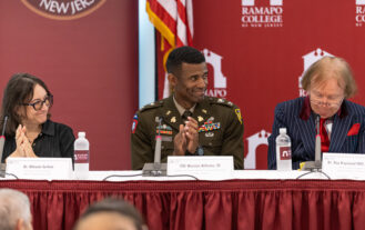 A group of three people sit at a table. The one in the middle has a military uniform on. There is an American flag behind them.