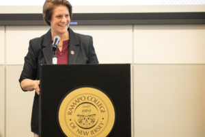 President Jebb is standing at a podium with the gold presidential seal on the front of it. She is smiling and looking to her left at someone in the crows (off-camera)