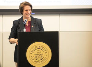 President Jebb is standing at a podium with the gold presidential seal on the front of it. She is smiling and looking to her left at someone in the crows (off-camera)