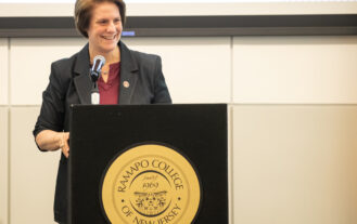 President Jebb is standing at a podium with the gold presidential seal on the front of it. She is smiling and looking to her left at someone in the crows (off-camera)