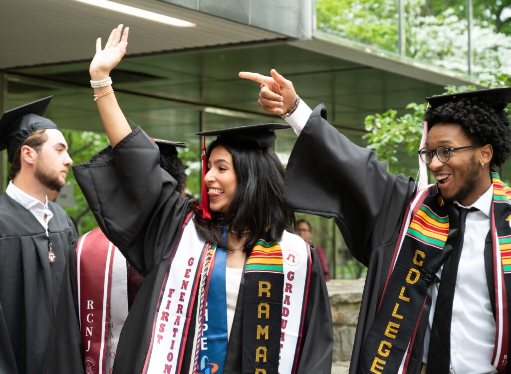 Two students in graduation caps and gowns are smiling and waving to their right off-camera. The caps and gowns are black, and both are wearing multiple stoles and cords, including ones that are for First Generation and for EOF. 