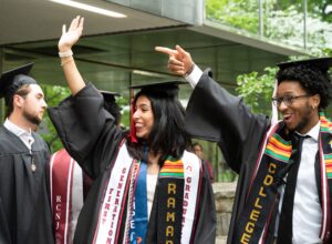 Two students in graduation caps and gowns are smiling and waving to their right off-camera. The caps and gowns are black, and both are wearing multiple stoles and cords, including ones that are for First Generation and for EOF.