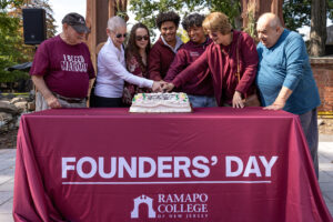 A group of 7 people stand behind a Ramapo College of New Jersey table outside. They cut a birthday cake.
