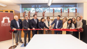 11 people stand behind a red ribbon in a lab. The person in the middle holds scissors to the ribbon.