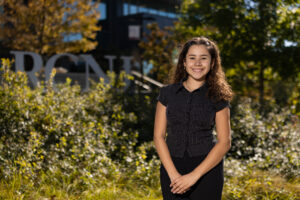 A young woman stands outside in front on long grass and an RCNJ sign.