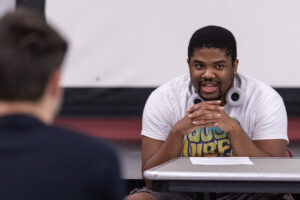 A young man sits at a table, with his hands folded below his face. He wears headphones around his neck and speaks.