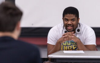 A young man sits at a table, with his hands folded below his face. He wears headphones around his neck and speaks.