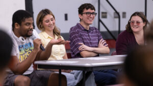 A group of four college students sit behind a table and laugh. The young man on the left holds a microphone and speaks.