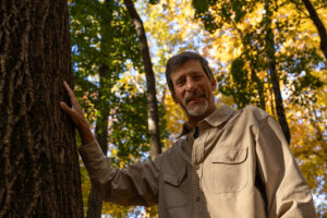 A man stands with his hand on a large tree. Yellow and green leaves are behind him.