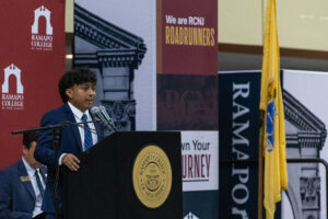 A young man stands at a podium with a Ramapo College seal. Ramapo College posters are behind him.