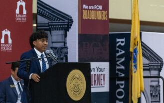 A young man stands at a podium with a Ramapo College seal. Ramapo College posters are behind him.