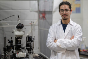 A young man wearing a white lab coat stands with his arms crossed in a lab next to a large microscope.