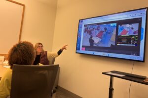A woman sitting at a table points to a television screen on the wall of a room. A person is sitting in a chair across from her and looks at the screen. The screen shows a moch hospital room and nursing students.