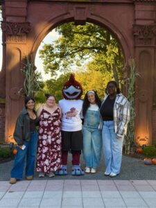 A Rocky the Roadrunner mascot is flanked by four people outside in front of an arch.