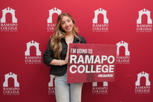 A young woman smiles and holds a sign that reads, I'm Going to Ramapo College. She is front of a maroon Ramapo College backdrop.