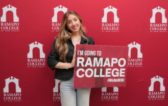 A young woman smiles and holds a sign that reads, I'm Going to Ramapo College. She is front of a maroon Ramapo College backdrop.