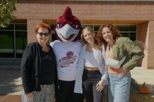Three generations of women stand next to a Rocky the Roadrunner mascot . The youngest is a college student. They are all outside.