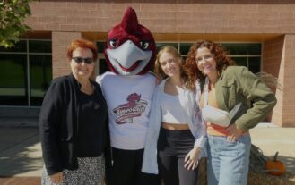 Three generations of women stand next to a Rocky the Roadrunner mascot . The youngest is a college student. They are all outside.