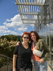 Two women stand outside with their arms around eachother. There are mountain views behind them, and they are next to a glass building.