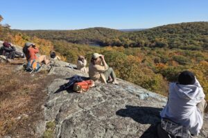 College students sit on a large rock and look out over the wooded mountains.