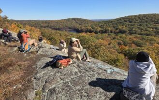 College students sit on a large rock and look out over the wooded mountains.