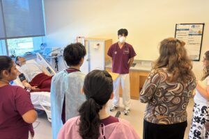 A nursing student stands near a hospital bed with a mannequin laying in it and talks to other college students in front of him.