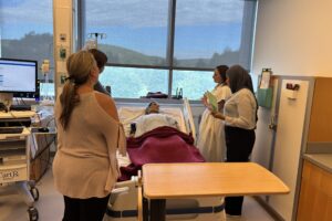 Four college students stand around a mannequin laying in a hospital bed. A large window with mountain veiws is behind the hospital bed.