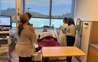 Four college students stand around a mannequin laying in a hospital bed. A large window with mountain veiws is behind the hospital bed.
