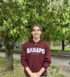Sebastian in a maroon Ramapo College hoodie stands smiling with hands clasped in front, outdoors near a tree with green leaves and a grassy area.