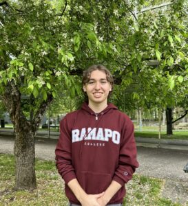 Sebastian in a maroon Ramapo College hoodie stands smiling with hands clasped in front, outdoors near a tree with green leaves and a grassy area.