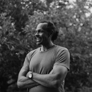 Person standing outside by the trees with arms crossed, smiling.