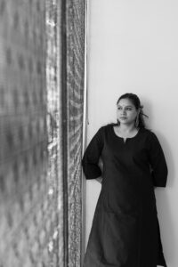 A woman in a long, dark dress stands indoors, leaning against a wall with her hands behind her back, looking thoughtfully into the distance; the image is in black and white.