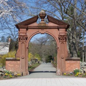 Ramapo College's Havemeyer Arch stands over a pathway lined with yellow flowers and trees, under a clear blue sky.