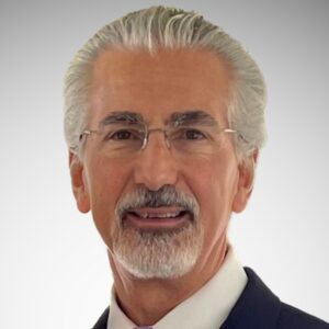 A man with white hair and a neatly trimmed white beard and mustache, wearing glasses and a dark suit with a white shirt, poses against a light gray background.