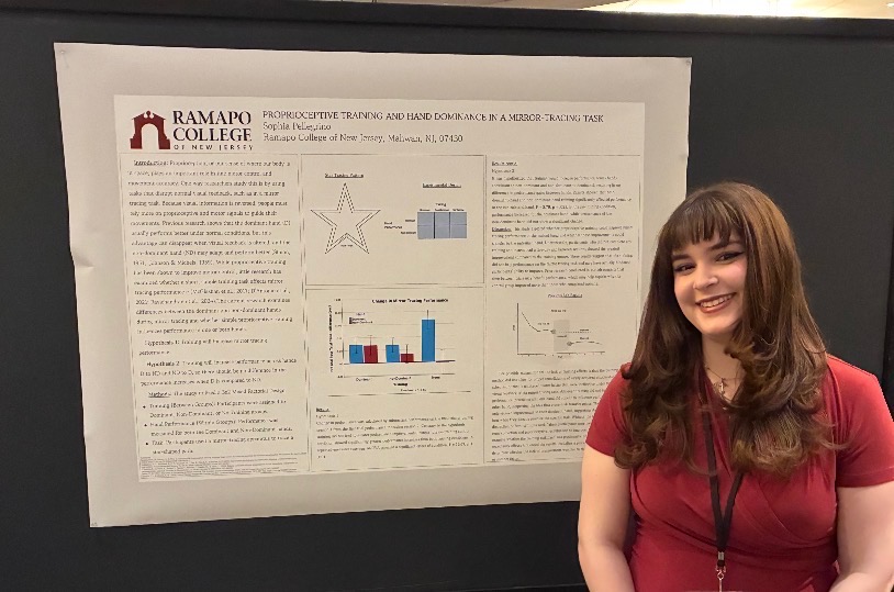 A woman with brown hair, wearing a red shirt and black lanyard, stands smiling in front of a scientific research poster from Ramapo College of New Jersey displayed on a black board.